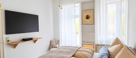 A bedroom with a large bed, a ceiling fan, a mounted TV, and a window with sheer curtains.