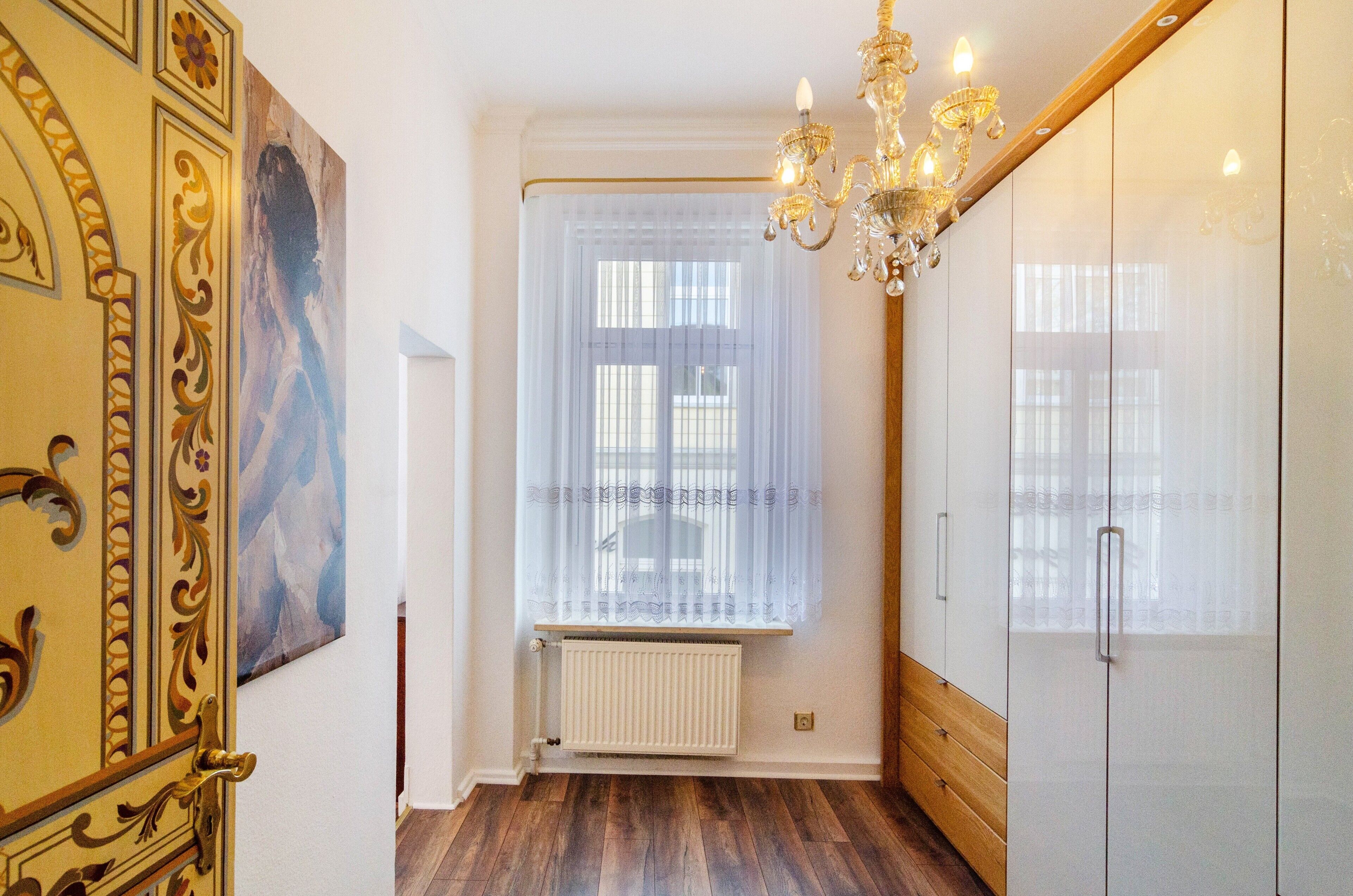 A room with a radiator, a window with sheer curtains, a chandelier, and a door with ornate design.