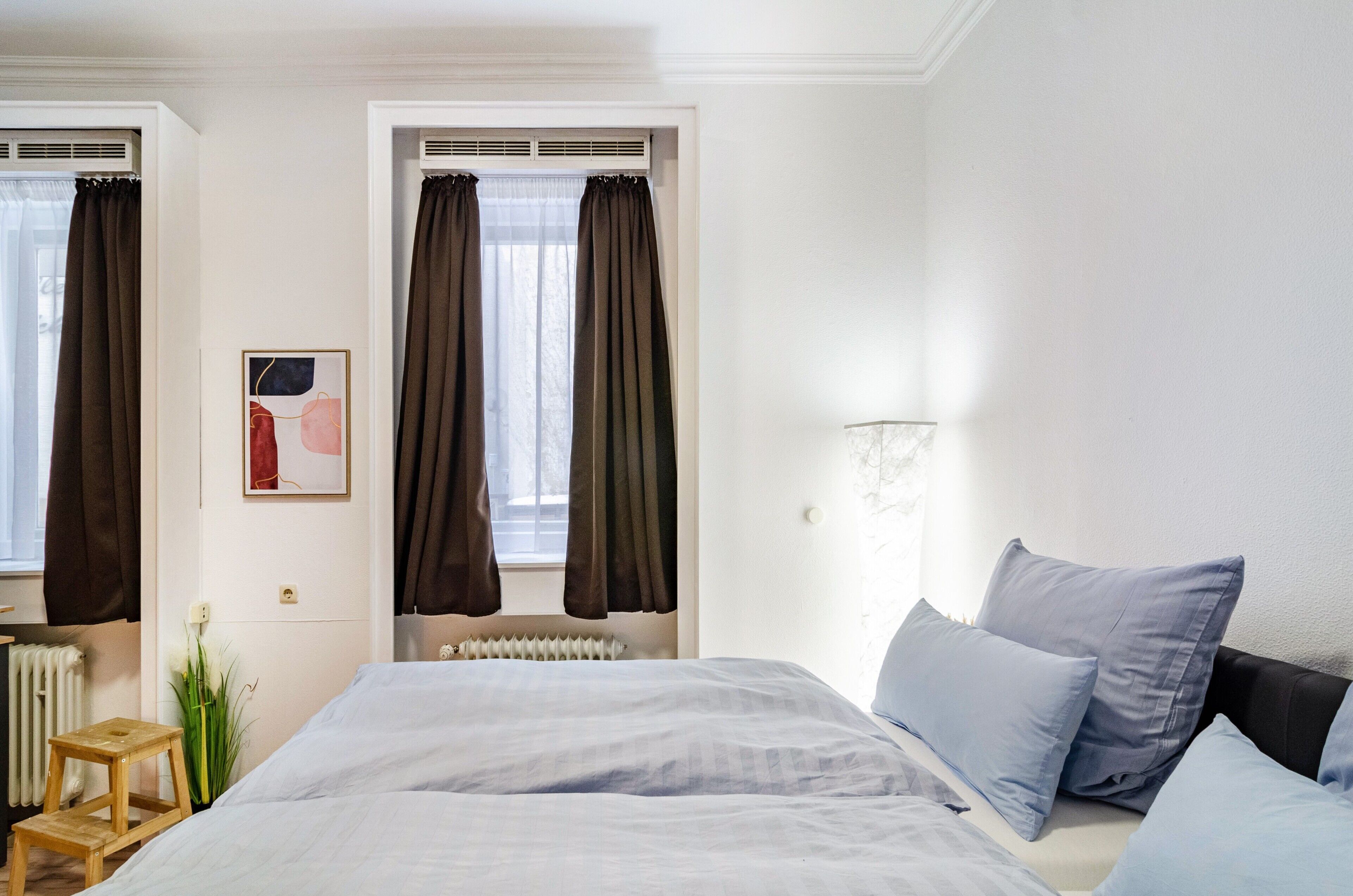 A bedroom with a bed, two windows with curtains, a radiator, a small wooden stool, and a wall-mounted light fixture.