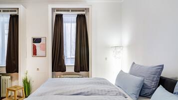 A bedroom with a bed, two windows with curtains, a radiator, a small wooden stool, and a wall-mounted light fixture.