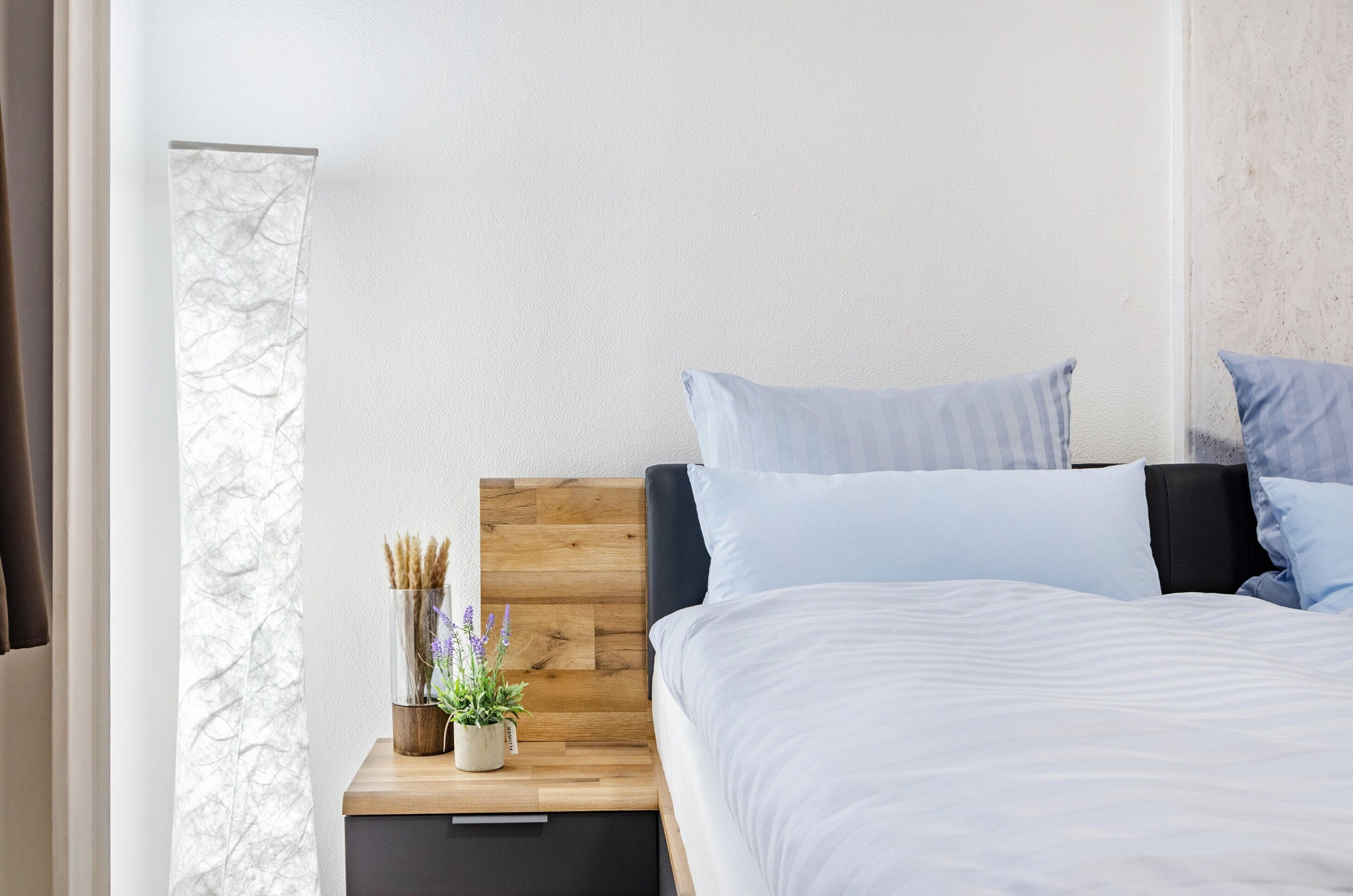 A bedroom with a wooden headboard, a bedside table with a plant, and a bed with white bedding.