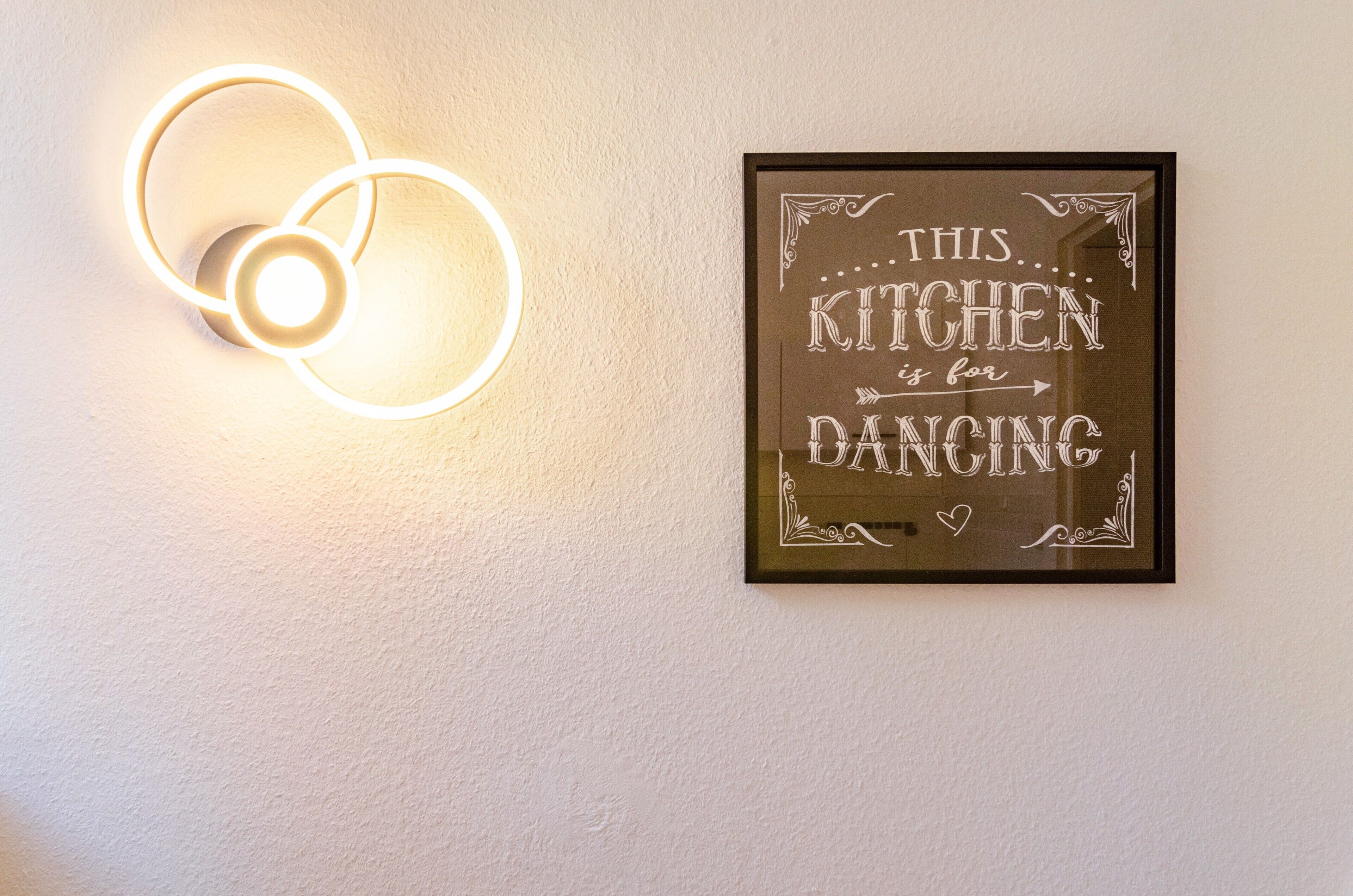Eine Wand mit einem gerahmten Schild, auf dem ‘THIS KITCHEN is for DANCING’ steht, und einer runden Leuchte.