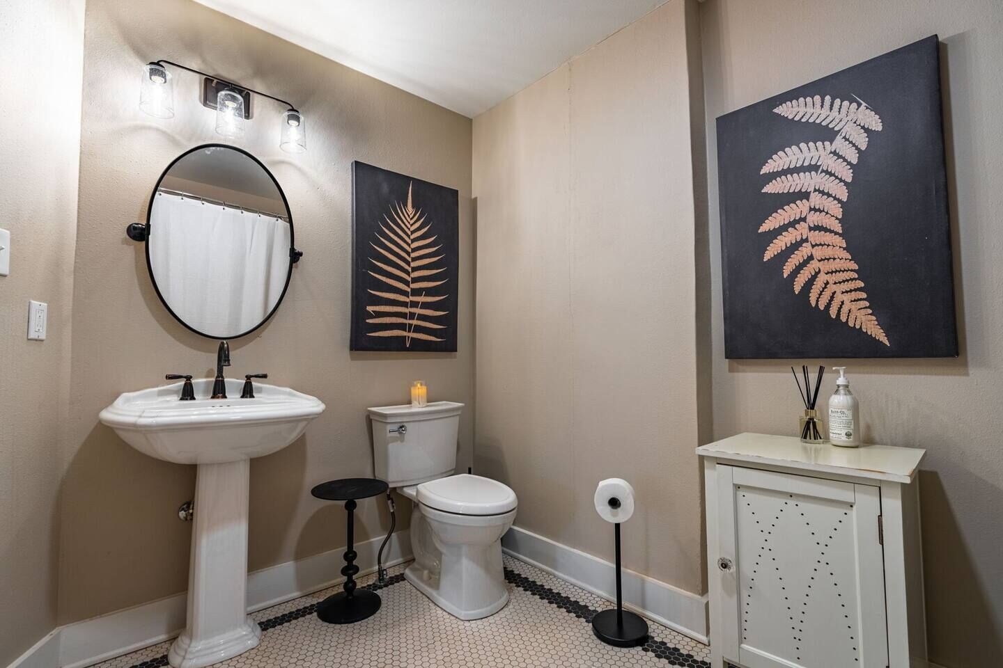 Une salle de bain avec un lavabo blanc, des toilettes, un bidet et un meuble de rangement. Il y a deux lampes fixées au mur et deux motifs de feuilles encadrées.
