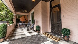 Een overdekt terras met een geruit vloertje, twee stoelen, een plafondventilator en een deur met een decoratieve ijzeren poort.