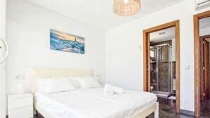 Ein Schlafzimmer mit Bett, Nachttisch, einem Gemälde, einem Badezimmer mit Dusche und einer Tür zu einem weiteren Raum.