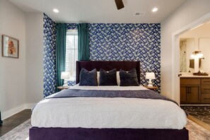 Un dormitorio con una cama grande, una mesita de noche con lámpara, una ventana con cortinas y un baño visible a través de una puerta abierta.