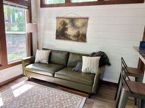 A room with a green leather sofa, a patterned rug, a painting on the wall, and a window with a view of trees. - Peaceful tiny home in cozy mentone (Mentone)