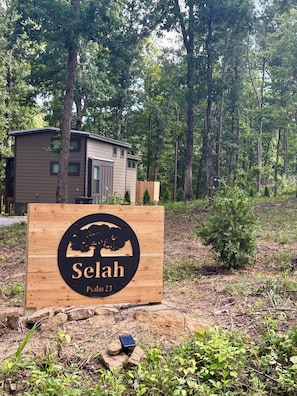 A wooden sign with a tree silhouette and the text 'Selah Psalm 23' in a forested area with a modern building in the background. - Peaceful tiny home in cozy mentone (Mentone)