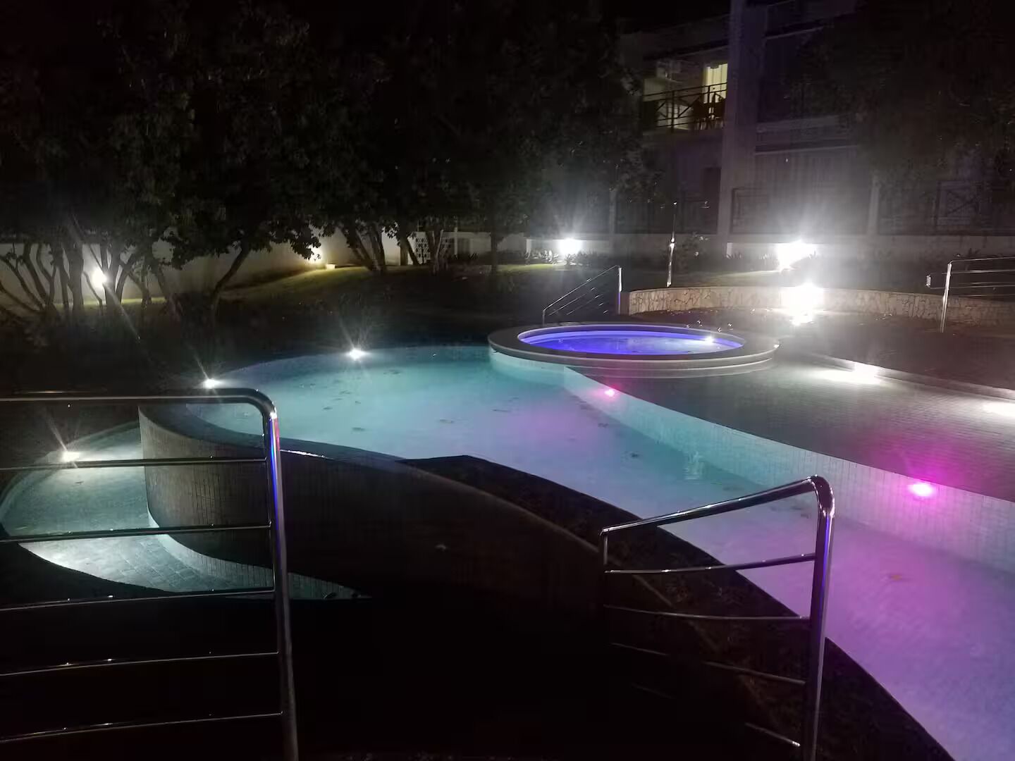 Ein Schwimmbad mit Whirlpool, umgeben von Bäumen und einem Gebäude im Hintergrund.