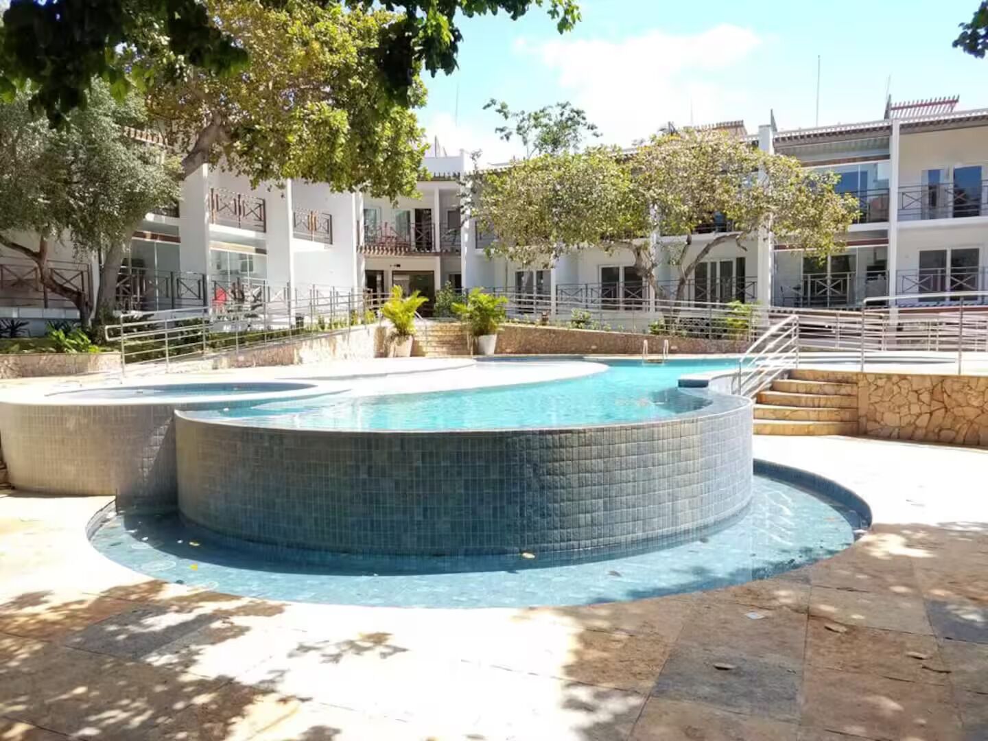 A swimming pool with a tiled edge, surrounded by a stone deck, and adjacent to a building with a balcony.