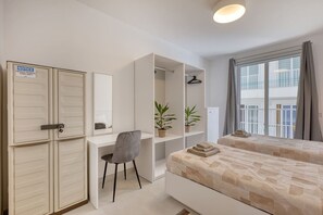 Ein Hotelzimmer mit Schreibtisch, zwei Betten, einem Kleiderschrank und einem Fenster mit Blick auf Gebäude.