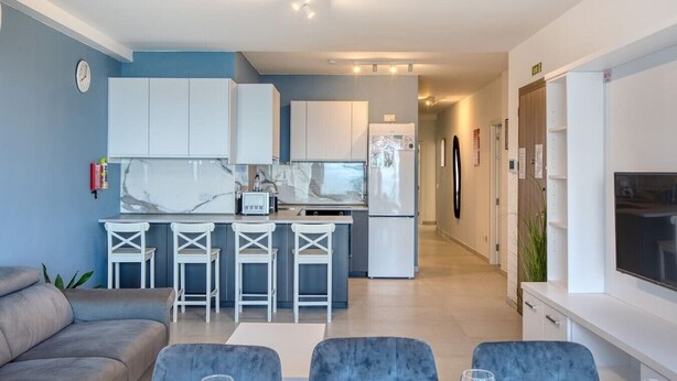 Een moderne woonkamer met een bank en fauteuils, een keuken met witte kastjes en een marmeren aanrechtblad, en een aan de muur bevestigde televisie.