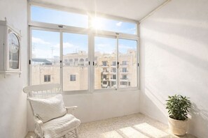 A balcony with a white chair, a potted plant, and a view of buildings through large windows. - The Elegant Hyacinth Flat (St. Paul`s Bay)