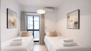 Ett hotellrum med två sängar, ett stort fönster med gardiner, en takfläkt och två inramade konstverk på väggarna.