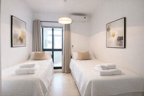 Ein Hotelzimmer mit zwei Betten, einem großen Fenster mit Vorhängen, einem Deckenventilator und zwei gerahmten Kunstwerken an den Wänden.
