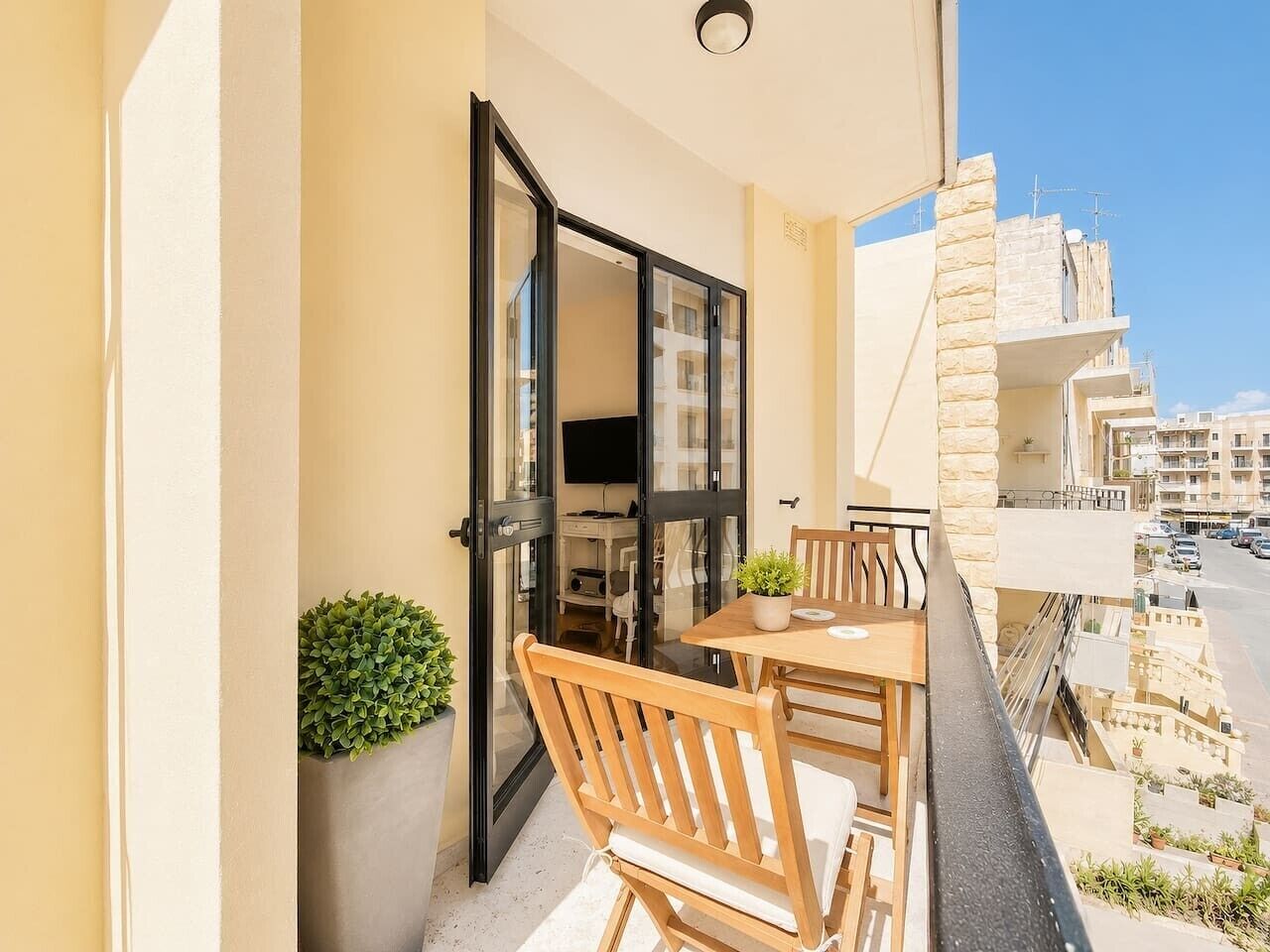A balcony with a table and chairs, a potted plant, and a view of the street and other buildings.