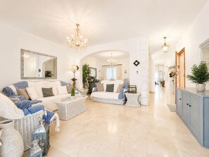 Ein geräumiges Wohnzimmer mit einer Couch, Sesseln und einem Couchtisch. Es gibt eine Chandelier, einen Spiegel und ein Sideboard.