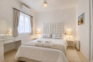 A hotel room with a bed, bedside tables, a mirror, a window with curtains, an air conditioning unit, a chandelier, and a painting on the wall. - The Elegant Hyacinth Flat (St. Paul`s Bay)