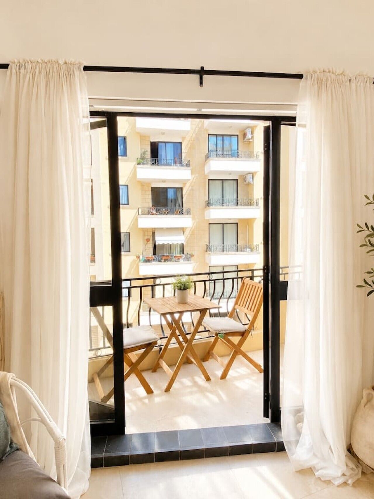 A balcony with a table and chairs, a view of a building with balconies, and sheer curtains.