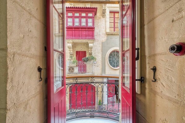 Ein schmaler Gang mit einer roter Tür, der zu einem Balkon mit schmiedeeisernem Geländer und Blick auf ein Gebäude mit roten Fensterläden führt.