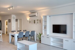 A modern living room with a dining area, a flat-screen TV mounted on a white entertainment unit, and a potted plant. - Modern Apartment with Sea View Qawra (St. Paul`s Bay)