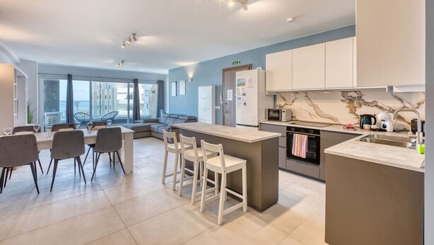 A modern kitchen with a central island, bar stools, and a dining area with chairs. - Modern Apartment with Sea View Qawra (St. Paul`s Bay)