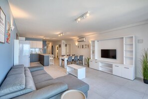 A modern living room with a grey sofa, a dining area with chairs, a kitchen in the background, and a TV on a white entertainment unit. - Modern Apartment with Sea View Qawra (St. Paul`s Bay)