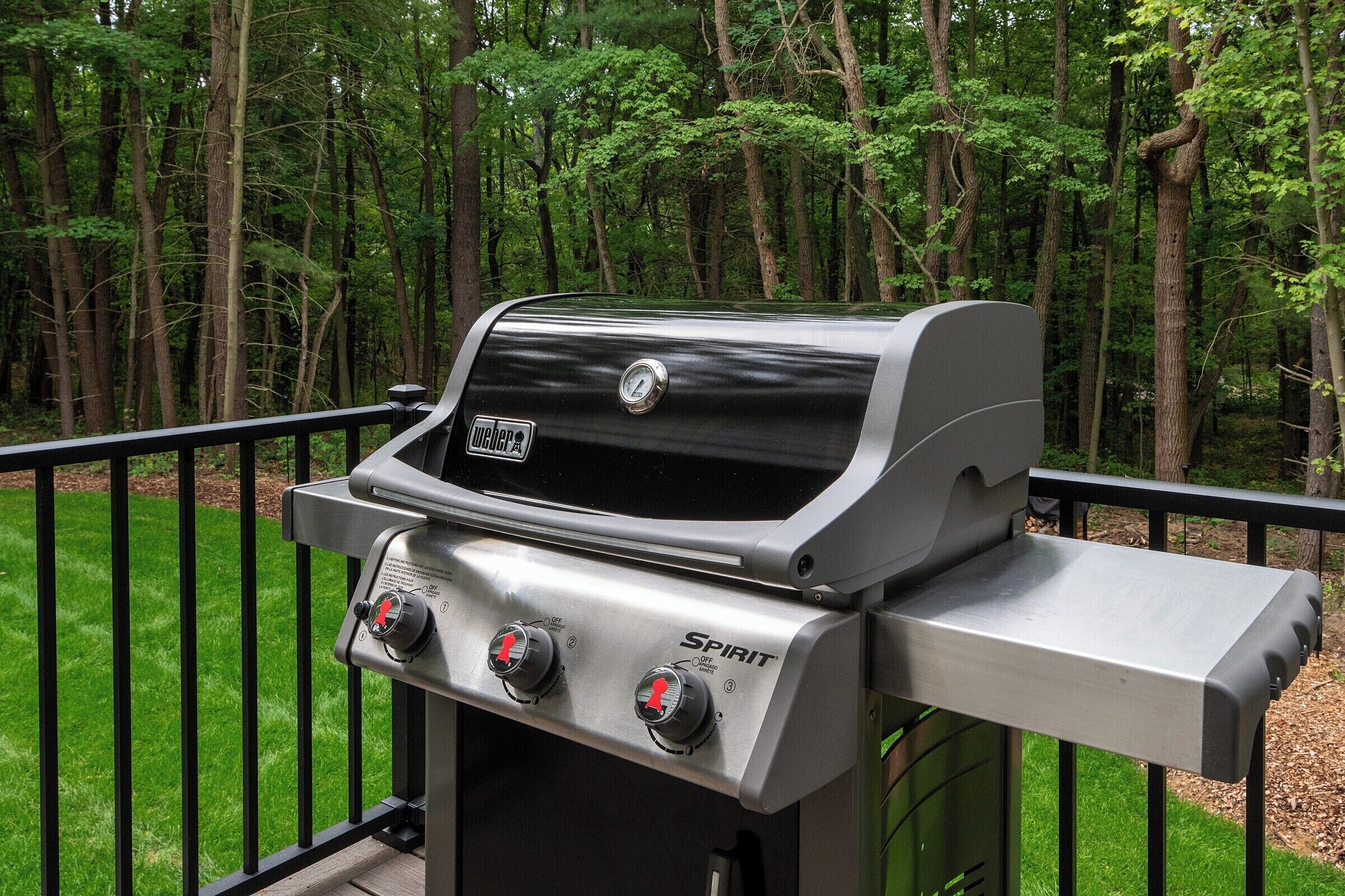 Una parrilla Weber Spirit con tres perillas de control, frente a un deck cubierto de pasto y un bosque.