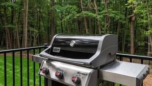 Una parrilla Weber Spirit con tres perillas de control, frente a un deck cubierto de pasto y un bosque.