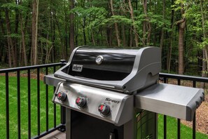 A Weber Spirit grill with three control knobs, set against a backdrop of a grassy deck and a forest. - The Dutch at Black Barn Ridge : Luxury Wellness in the Woods (Saugatuck)