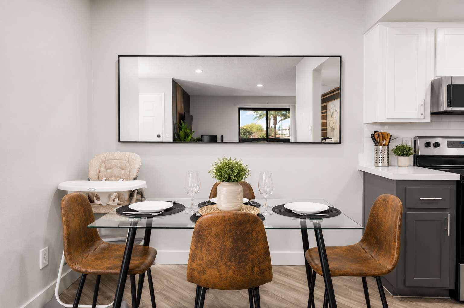 Un espace repas moderne avec une table en verre, deux chaises marron, une chaise blanche et un grand miroir au-dessus de la table.