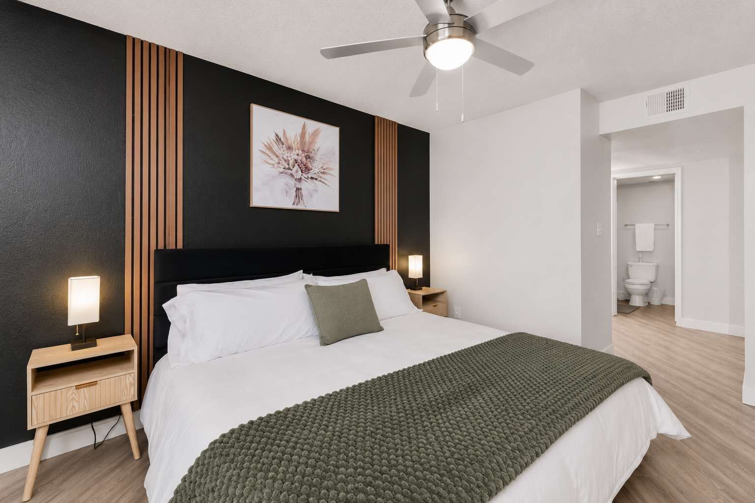 A modern hotel room with a large bed, a ceiling fan, a framed artwork on the wall, and a view into the bathroom.