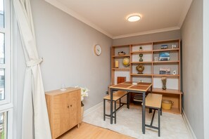 Une petite pièce comprenant un meuble en bois, une table avec deux chaises, une bibliothèque contenant divers objets et une fenêtre avec des rideaux.