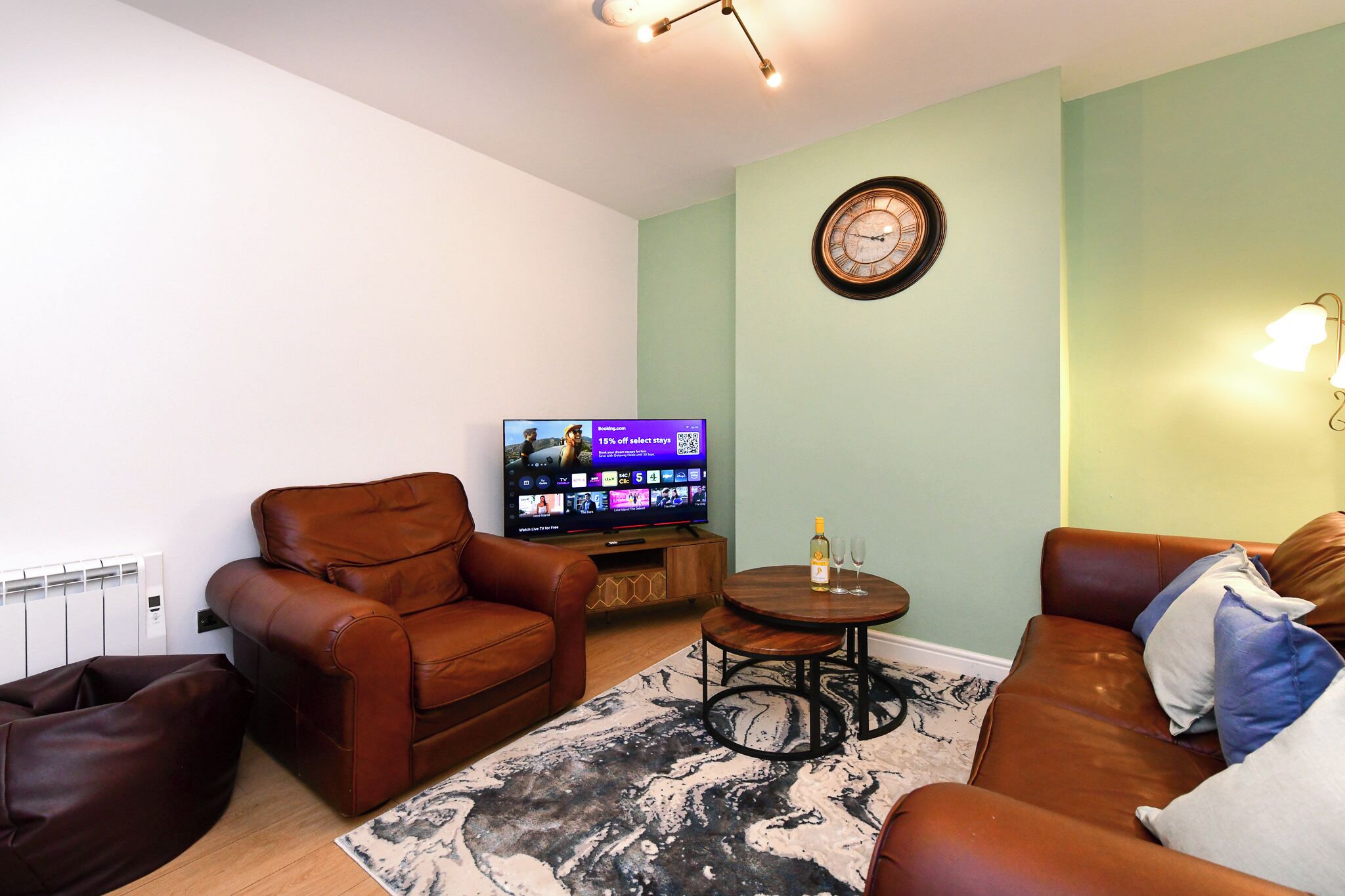 A living room with a brown leather sofa, a brown leather armchair, a small wooden coffee table, a flat-screen TV, and a decorative wall clock.
