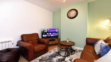 A living room with a brown leather sofa, a brown leather armchair, a small wooden coffee table, a flat-screen TV, and a decorative wall clock.