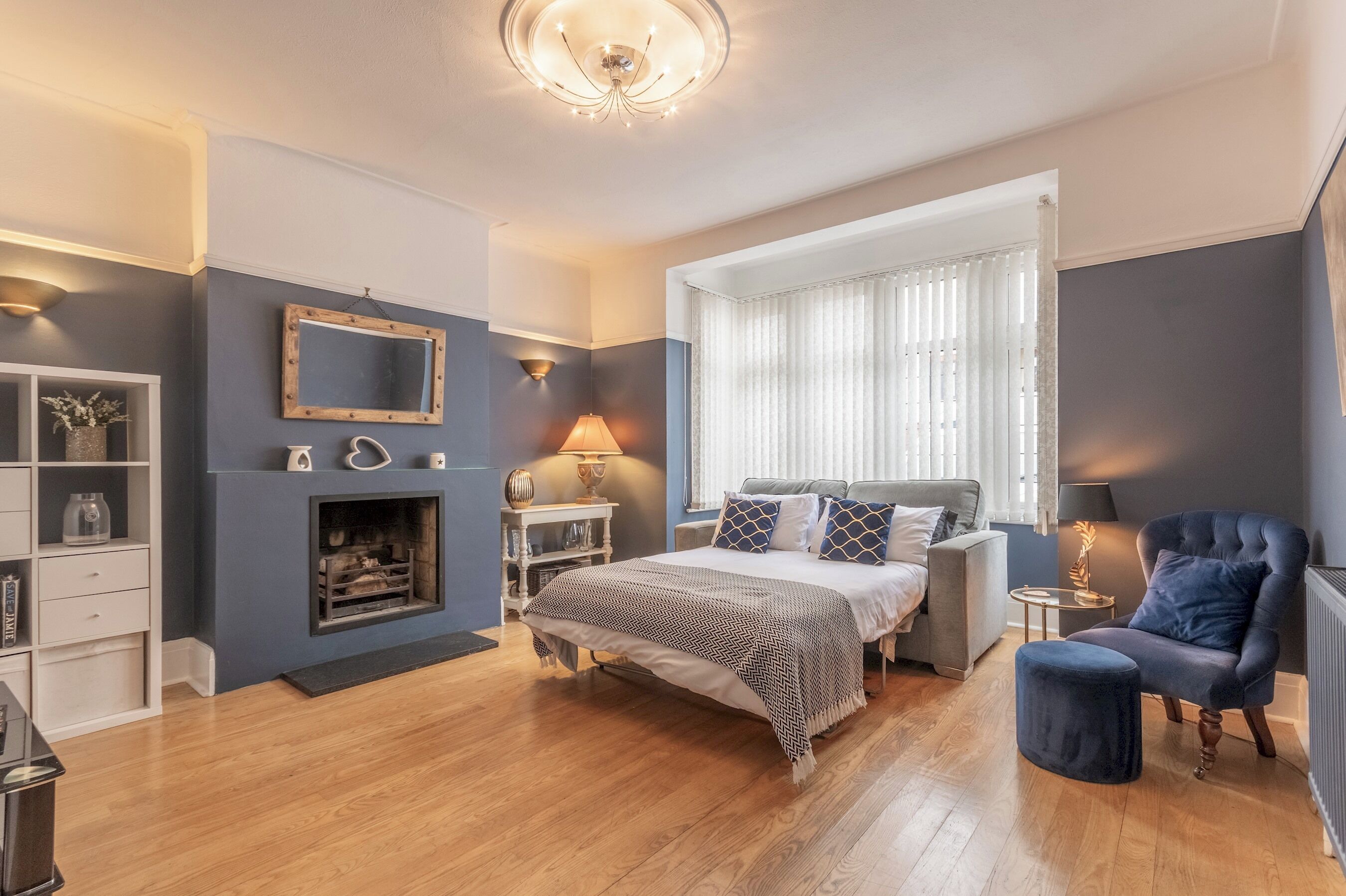 Une chambre à coucher avec un lit, des tables de chevet, un miroir, une cheminée, une fenêtre avec des rideaux et un fauteuil bleu.