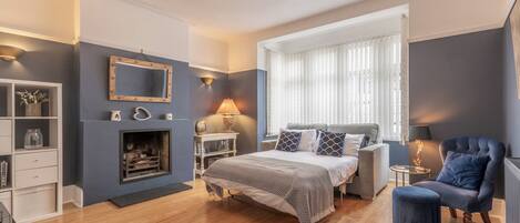 Un dormitorio con cama, mesitas de noche, un espejo, chimenea, ventana con cortinas y un sillón azul.