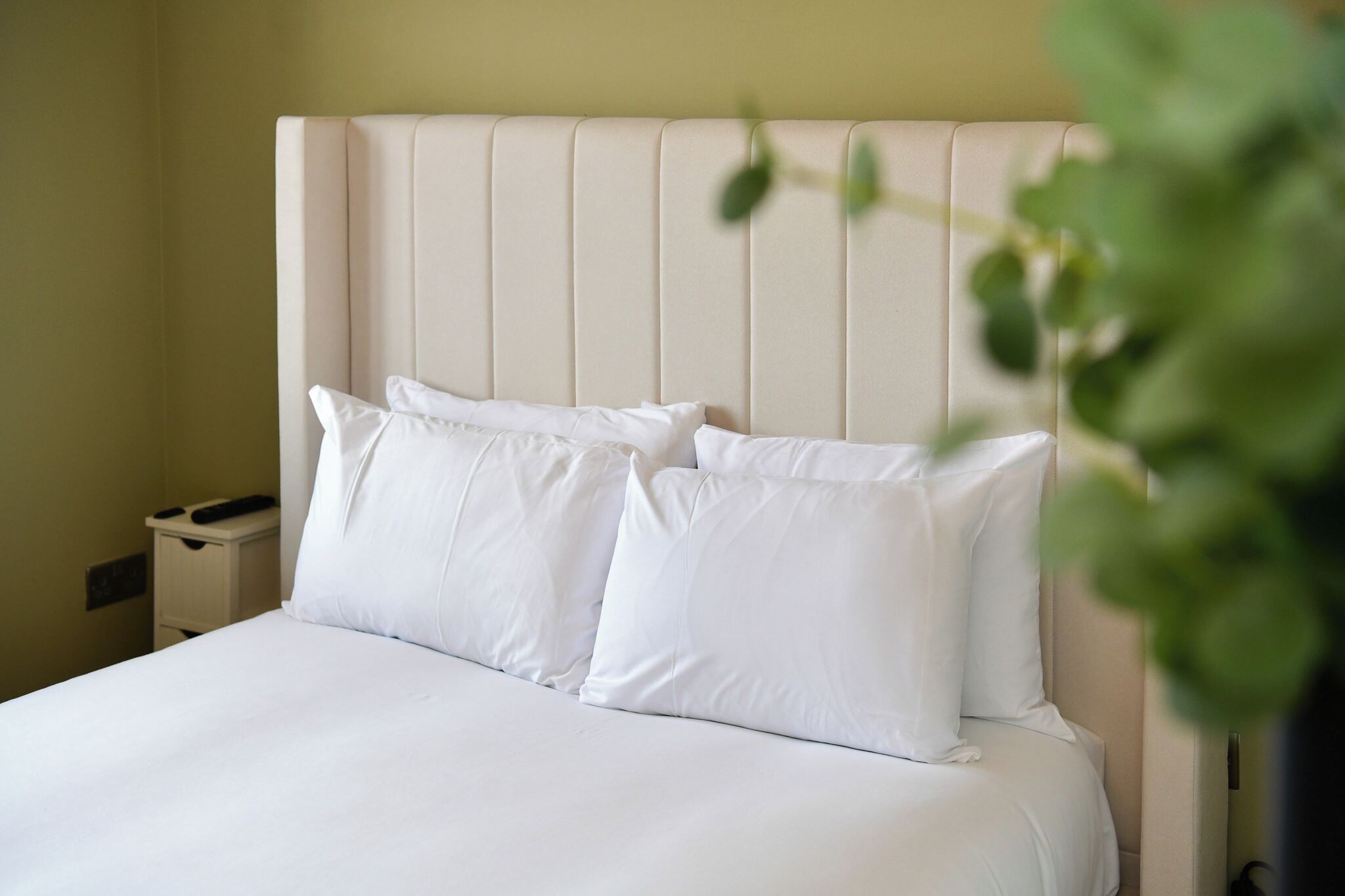 A neatly made bed with white linens and pillows, a bedside table with a lamp, and a wall-mounted switch.
