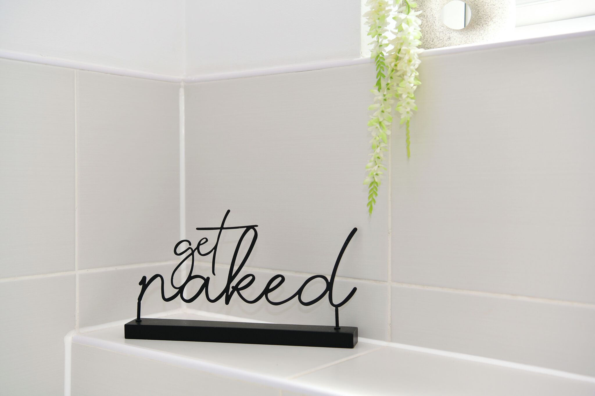 A bathroom with white tiles, a black metal sign with the word 'get naked,' and a hanging plant.