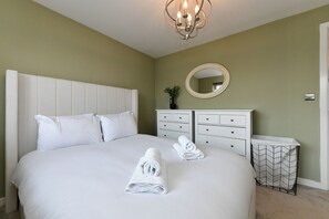 A neatly made bed with white linens and rolled towels, a white dresser with drawers, a round mirror on the wall, and a chevron-patterned laundry basket. - Stylish Family Home with Garden & Free Parking - Pass the Keys (Cardiff)