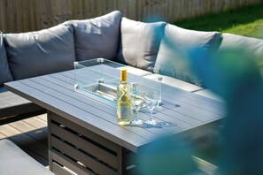 A glass-top table with a bottle of wine and two glasses, set on a patio with a cushioned seating area in the background. - Stylish Family Home with Garden & Free Parking - Pass the Keys (Cardiff)