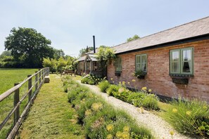 A brick building with green windows, a garden with various plants and flowers, and a wooden fence. - Charming Countryside Cottage with a Private Garden - Pass the Keys (Henley-in-Arden)