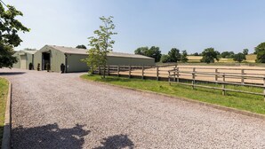 Ein Kiesweg führt zu einem Gebäude mit überdachter Einfahrt, einem eingezäunten Reitplatz und Bäumen im Hintergrund.