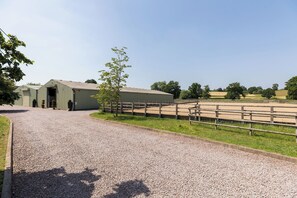 A gravel path leading to a building with a covered entrance, a fenced riding area, and trees in the background. - Charming Countryside Cottage with a Private Garden - Pass the Keys (Henley-in-Arden)