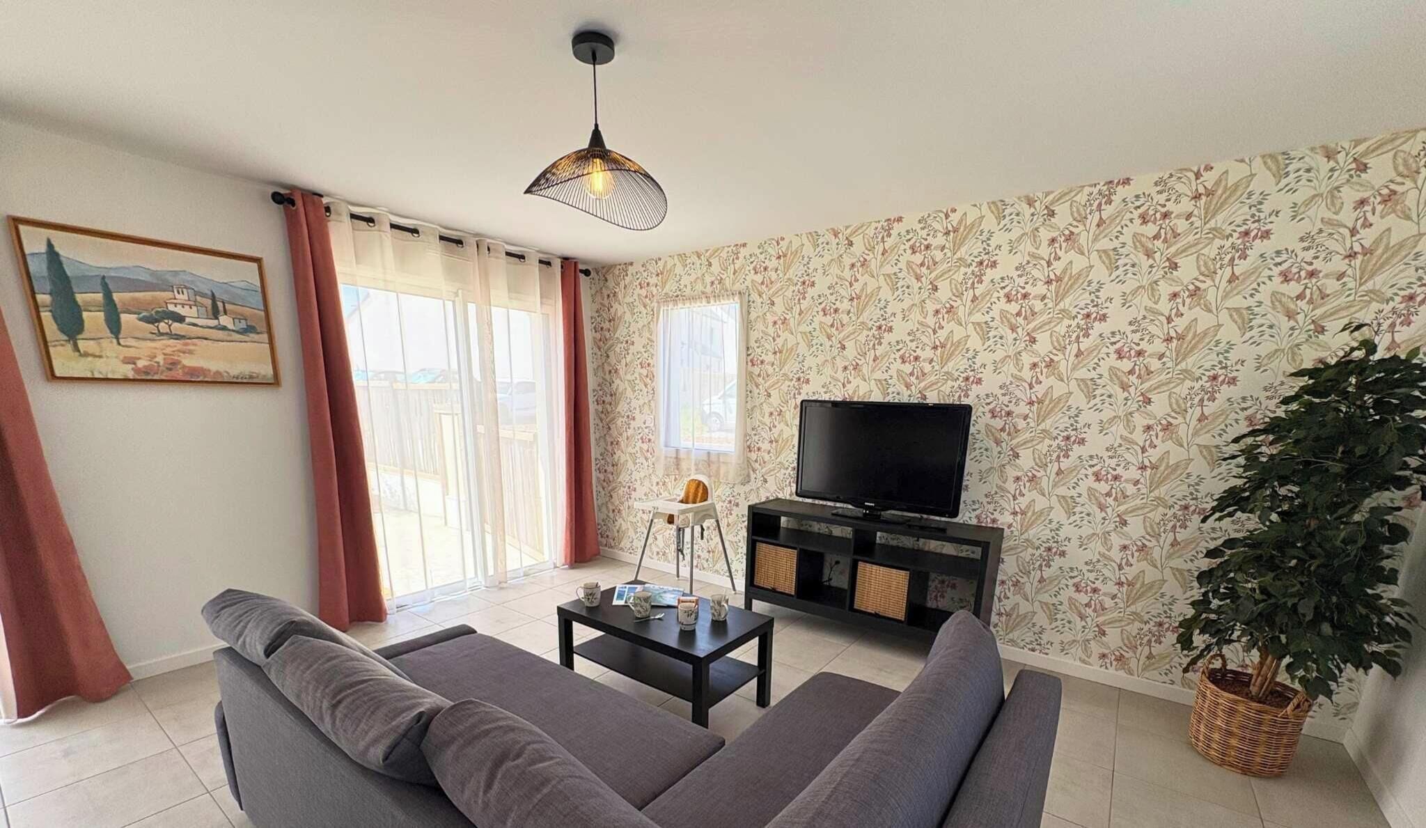 Ein Wohnzimmer mit einer Couch, einem Fernseher, einem Tisch mit Tassen, einem Gemälde, einem Fenster mit Vorhängen und einer Pflanze.