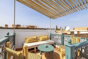 Eine Dachterrasse mit einer hölzernen Pergola, einem weißen Sofa mit gelben Kissen, einem runden Tisch und einem Korbstuhl.