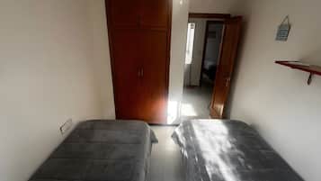 A room with two beds, a ceiling fan, a wooden wardrobe, and a small shelf.