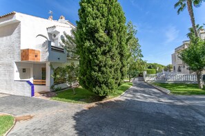 A residential street with a white house featuring a tiled roof, a large evergreen tree, and a paved driveway. - Holiday Home 'Playa, Piscinas Y Jardines' with Shared Pool, Wi-Fi and Air Conditioning (Alicante)