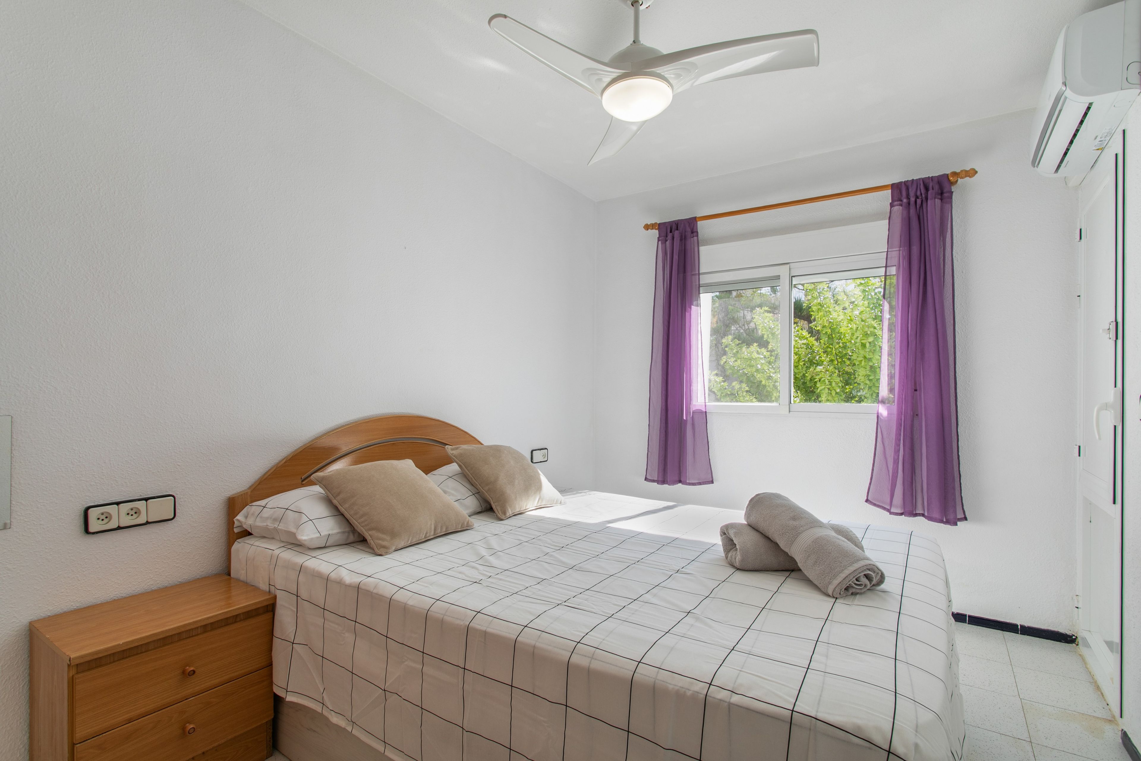 A bedroom with a bed, a wooden nightstand, a ceiling fan, a window with purple curtains, and an air conditioning unit.