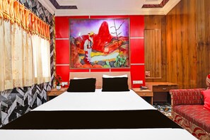 A hotel room with a bed, a nightstand, a desk, a chair, and a large painting on the wall. - Classic Hotel Bidisha (Kanthi)